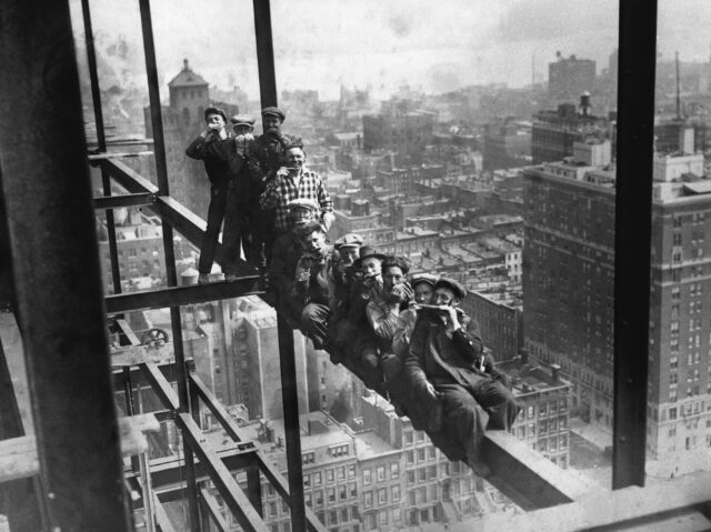 A line of construction workers eating lunch on a steel girder.