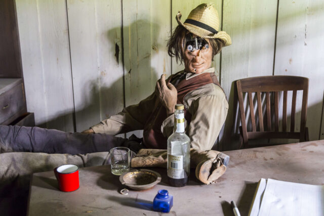 Animatronic dressed as a person in the Wild West sat in a chair, with various items in front of it on a wooden table