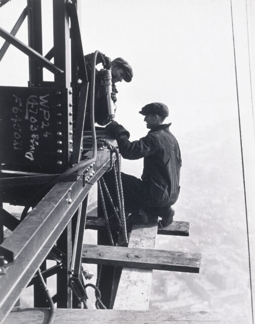 Two constructions workers at the corner of a skyscraper, working.