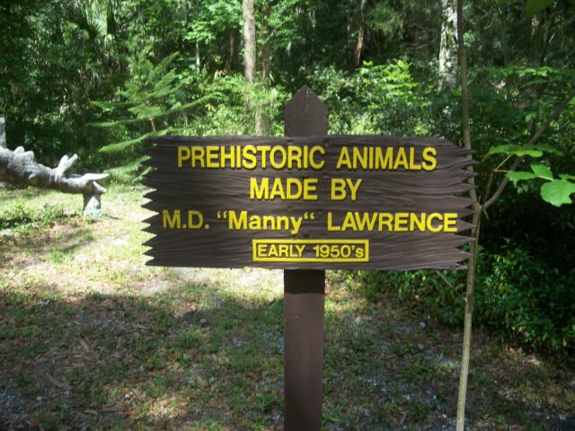 Sign reading "PREHISTORIC ANIMALS MADE BY M.D. 'Manny' LAWRENCE, EARLY 1950's" in the middle of a wooded area at Bongoland