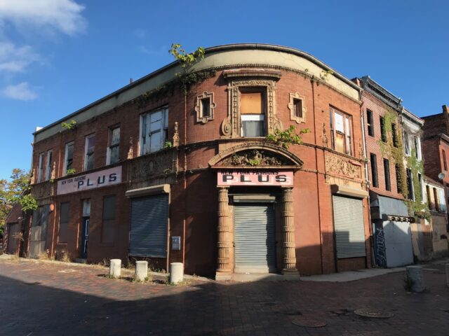 A corner building abandoned with plants growing up its walls.